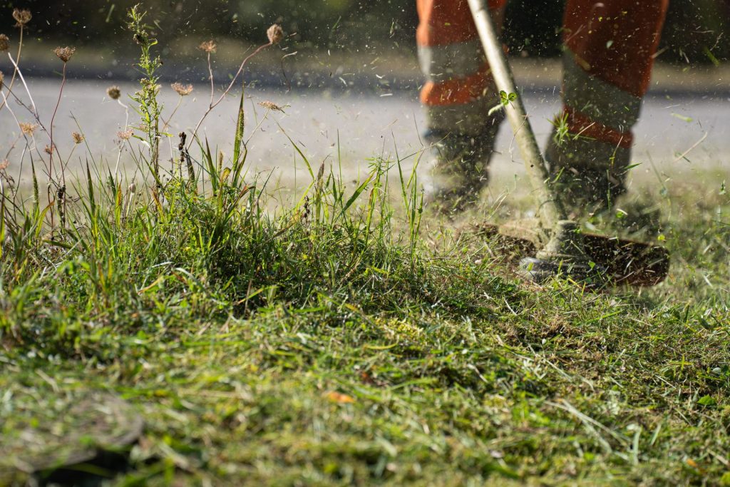 Ein Arbeiter in Schutzkleidung stutzt mit einem Rasentrimmer das Gras; die Aufnahme fängt die dynamische Bewegung und das umherfliegende Schnittgut ein.